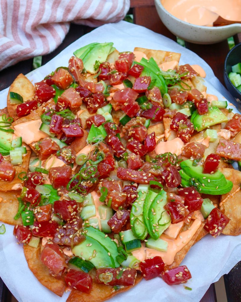 mejicano-poke-nachos-avocado-tuna-spicy-mayo A large plate of poke nachos is displayed on a light surface. The tortilla chips are generously topped with bright red seasoned tuna cubes, green avocado slices, sliced jalapeños, sesame seeds, and fresh cilantro leaves. An orange creamy sauce is also visible. In the blurred background, there's a bowl of diced cucumbers, lime wedges, and a glass of water, against a backdrop of lush greenery.