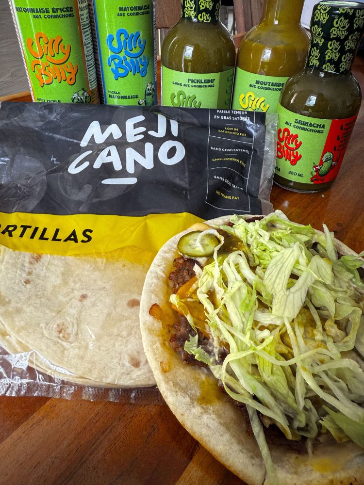 A top-down shot features an open taco with meat, melted cheese, shredded lettuce, and pickles on a tortilla, sitting on a Mejicano branded tortilla package. Surrounding it, several "Silly Billy" sauce bottles are visible in the background, including spicy pickle mayonnaise, pickle mustard, and pickle sriracha sauce, along with a bottle of pickle relish.