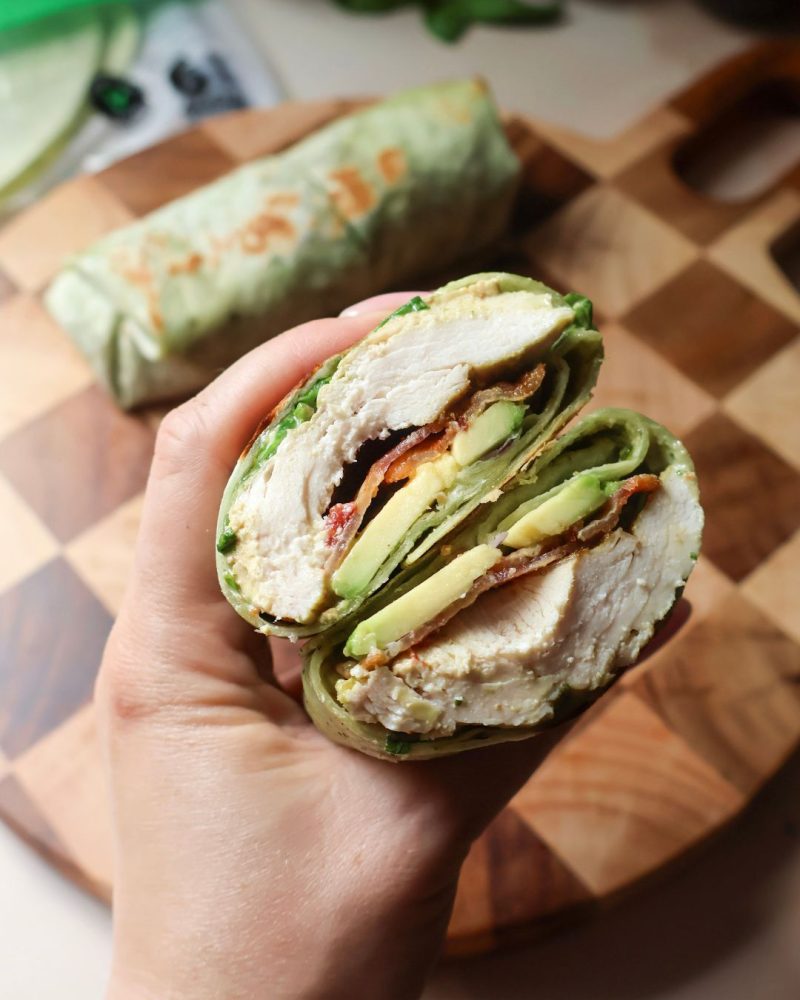 Un wrap coupé en deux garni de poulet grillé, bacon, avocat, roquette et sauce crémeuse au pesto, tenu à la main au-dessus d’une planche de bois. Un emballage de tortillas Mejicano est visible en arrière-plan.